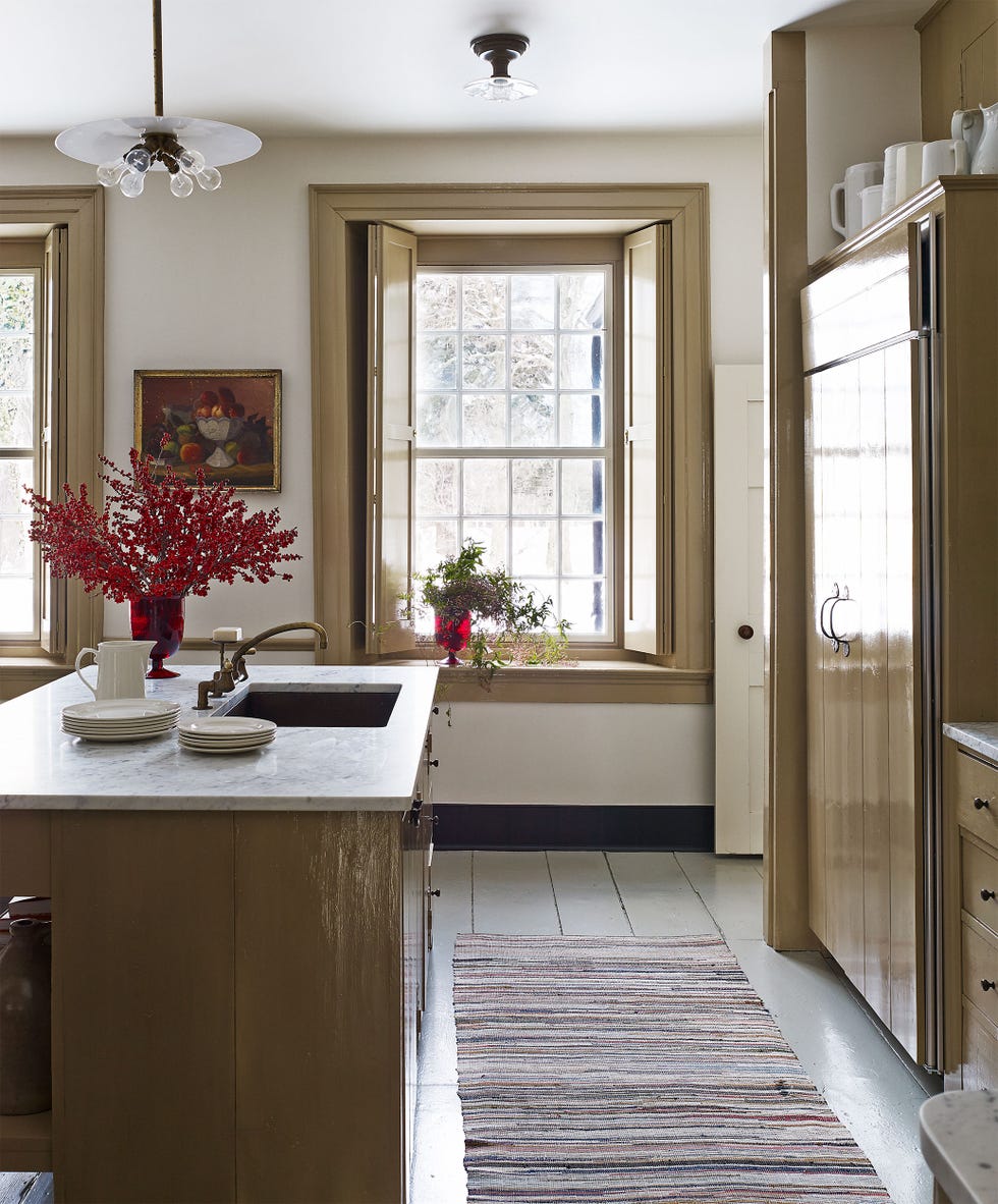 a modern kitchen features an island with a marble countertop and a sink there are several stacked white plates on the countertop a vibrant red vase with branches and a smaller vase with greenery add color and life bright light filters through the windows, which are opened to allow natural air in two large windows frame the view outside, and a painting hangs on the wall, contributing to the cozy atmosphere the cabinetry is designed in a warm, neutral palette, complemented by a patterned rug on the floor