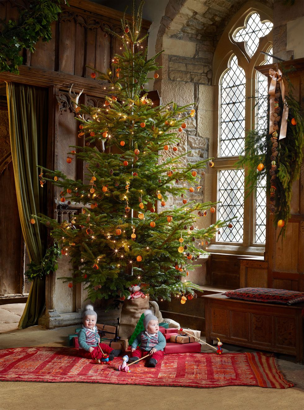 Twin children wearing blue sweaters with matching hats and pompoms sit next to a Christmas tree in an English country house