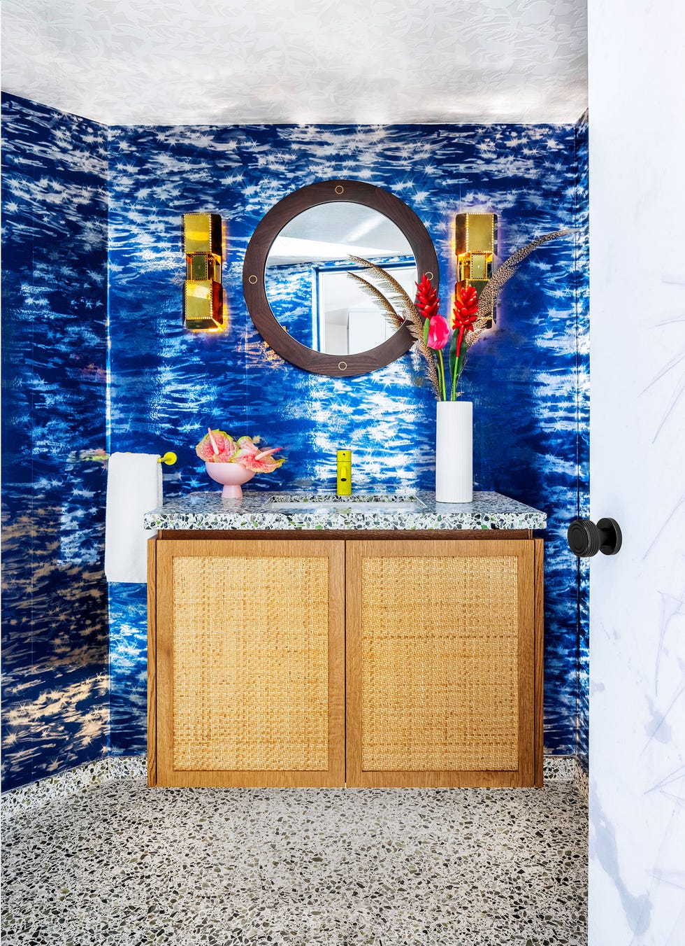 blue striated wallpaper in a powder room with rattan vanity and speckled marble sink oval mirror above it and a white vase with red flowers on top of the sink