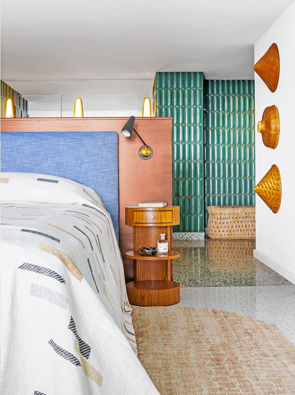bedroom with white cover with muted color stripes next to the bed is a round three tier night stand in wood and beyond are seen the green vertical tiles from the back of the bathroom wall
