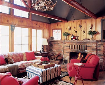 wood panelled living room with large brick fireplace and small mountains animal heads and a red pattenered cushy sofa and matching red leather chairs and a striped blue and white and gray ottoman at center with a tray with flowers and objects on it and antler chandelier above