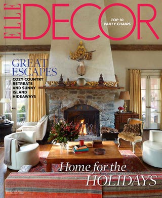 a spacious living room with a prominent stone fireplace as the focal point the fireplace features a decorative mantle adorned with ornate items to the left, a comfortable white sofa is positioned near a coffee table topped with books and a floral arrangement a vibrant area rug covers the wooden floor, complemented by various seating options, including an elegant armchair and plush furnishings large windows allow natural light to pour in, enhancing the warm atmosphere