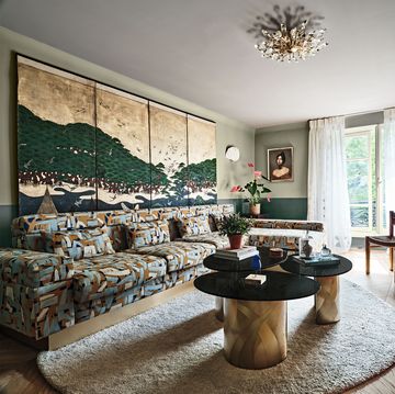 a living room with a large half moon rug, three pedestal cocktail tables with gold bases, plants, a mural behind the brown and blue geometric design sofa