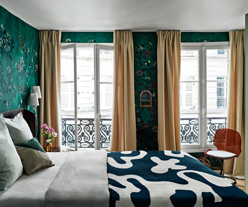 green patterned wallpaper on bedroom walls with skinny double doors looking out over a quintessential parisian balcony and a bed with a swirly pattern dark blue green blanket on top