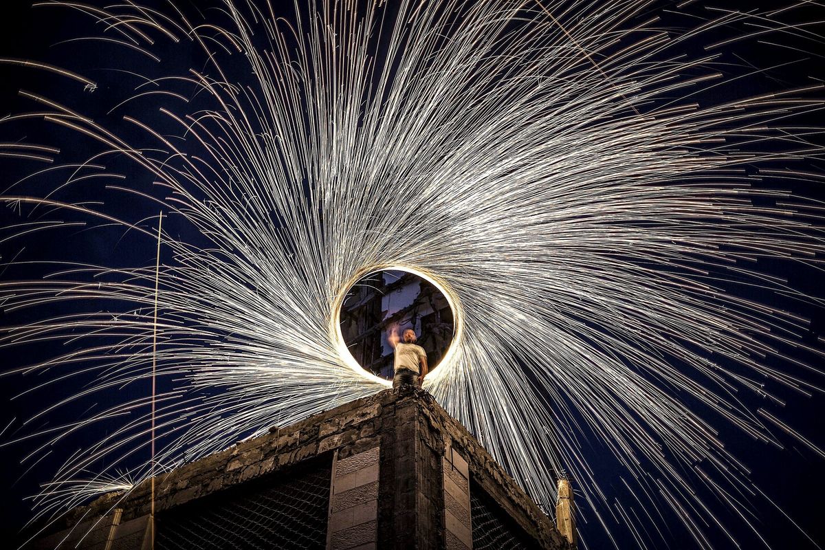Een man zwaait boven op een gebouw in GazaStad met vuurwerk waardoor hij een stralenkrans van licht maakt in de nachtelijke hemel