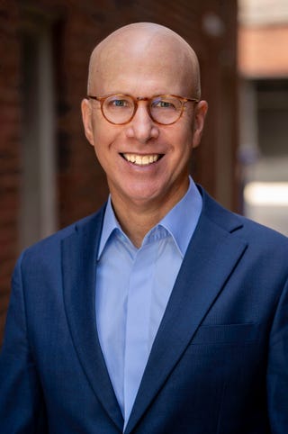 jonathan eig wearing a navy suit jacket and purple shirt, wearing glasses, smiling at the camera