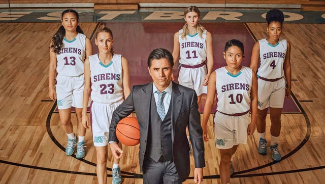imagen promocional de 'el míster', con los protagonistas mirando a cámara las actrices tiana le, sophia mitri schloss, cricket wampler, tisha custodio, monique green van vestidas con ropa de baloncesto delante, john stamos, de traje, sujeta una pelota de baloncesto
