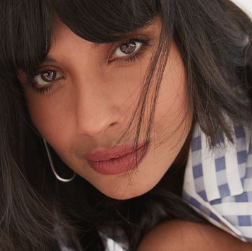 jameela jamil leans in toward the camera lens wearing a silver earring and a blue and white checked blazer