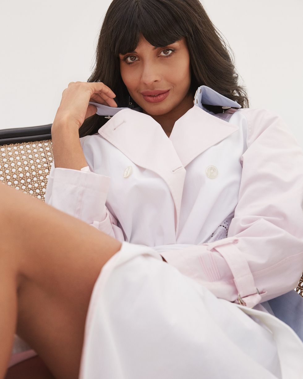 jameela jamil sitting on a chair against a white backdrop, wearing a pink trench coat by tommy hilfiger