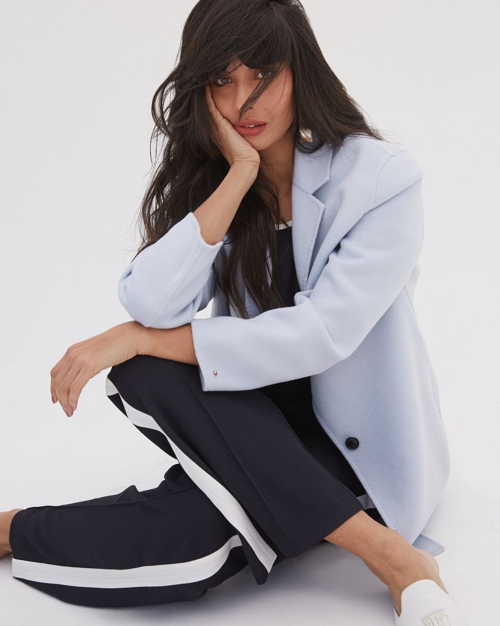 jameela jamil sits cross legged with her hair across her face, against a white backdrop, she is wearing a pale blue coat, navy trousers and white shoes