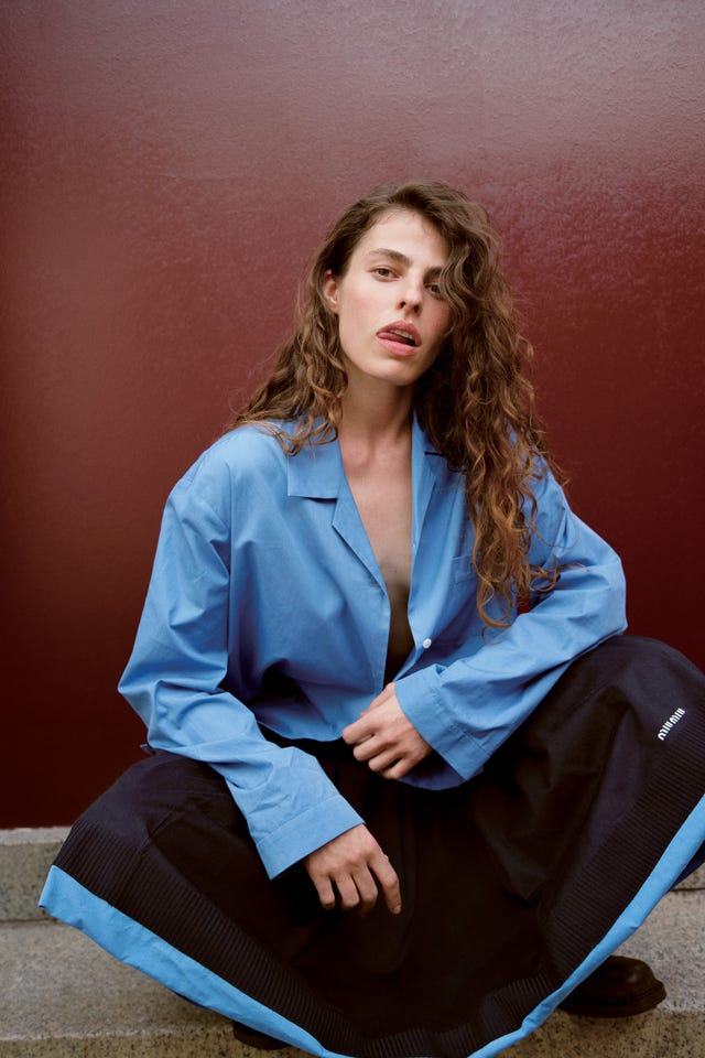 person wearing a blue shirt and a black skirt seated against a maroon wall