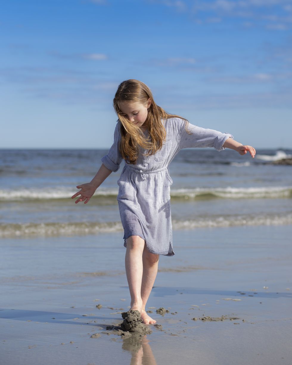 elementary age girl in blue dress walking on a beach and digging feet into the sand