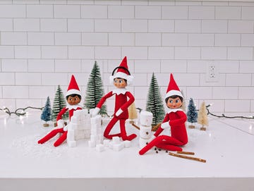 three elf on the shelf elves sit around making a snow fort out of sugar cubes and building a snowman out of marshmallows