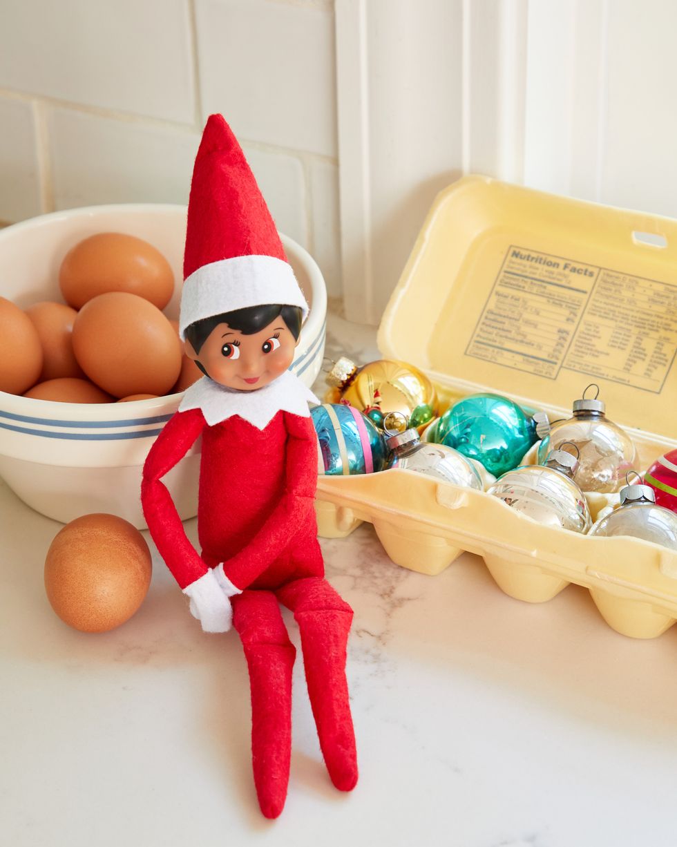 elf on the shelf sitting beside bowl of eggs and an open egg carton filled with christmas ornaments