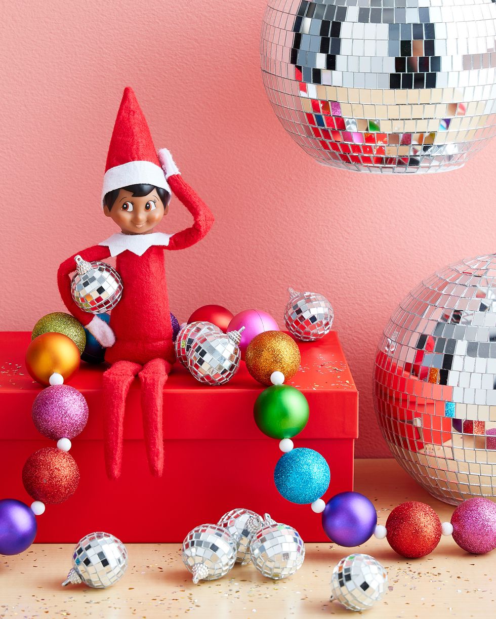 the elf on the shelf sitting on a red box with colorful beaded garland and tiny disco ball ornaments