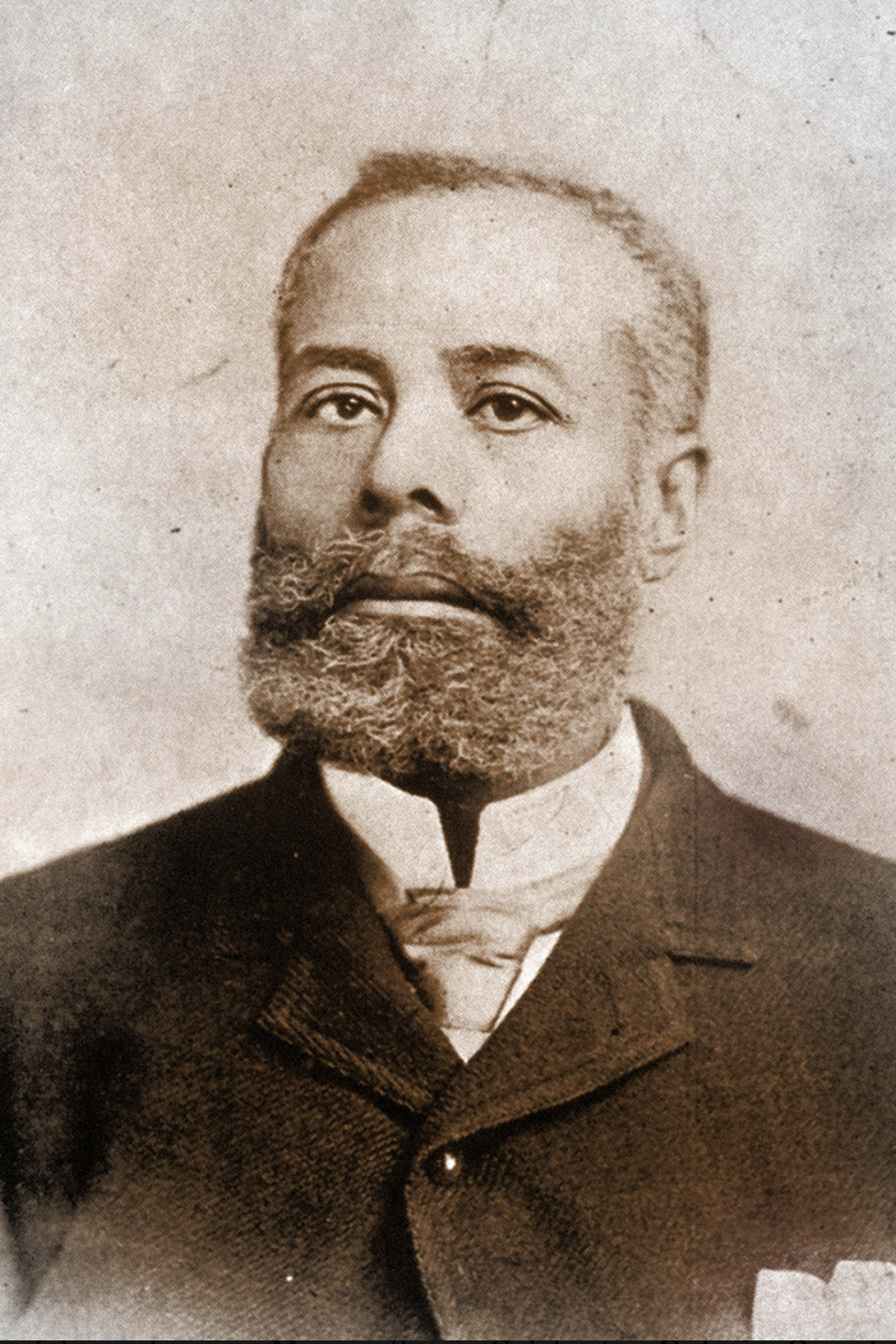 elijah mccoy looks at the camera, he wears a jacket over a collared shirt and tie and has a full beard