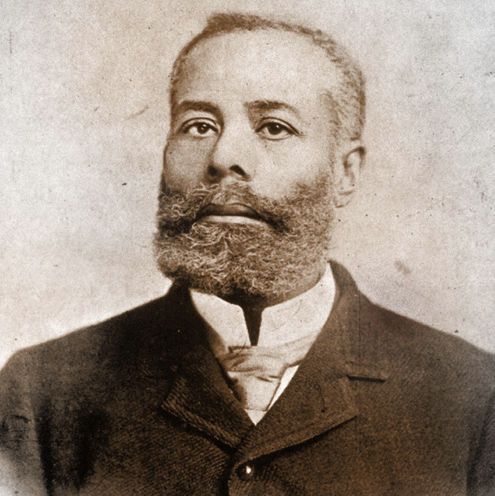 elijah mccoy looks at the camera, he wears a jacket over a collared shirt and tie and has a full beard