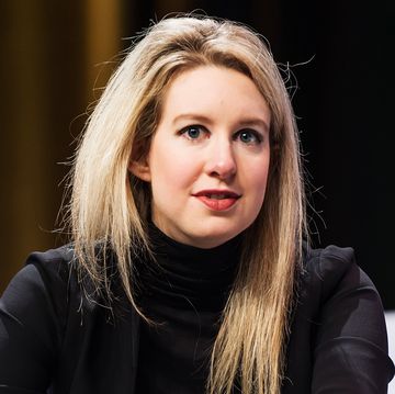elizabeth holmes wears a signature black turtleneck and red lipstick, her blonde hair is styled down, and she looks past the camera with a neutral expression on her face