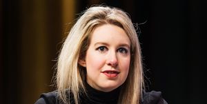 elizabeth holmes wears a signature black turtleneck and red lipstick, her blonde hair is styled down, and she looks past the camera with a neutral expression on her face