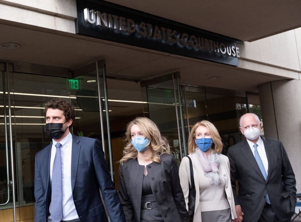 billy evans, elizabeth holmes, noel holmes, and christian holmes walk away from the doors of a united states courthouse, all four are wearing face masks and professional clothing, they look left of the camera and hold hands
