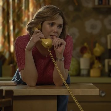 a photo from the miniseries love and death, with actress elizabeth olsen wearing a pink shirt and standing in a kitchen, leaning on the counter and speaking into a telephone receiver