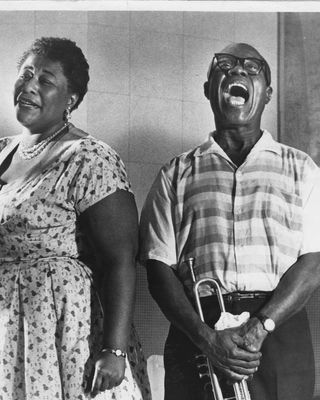 ella fitzgerald smiles and looks at the camera while wearing a short sleeve patterned dress, louis armstrong stands to the right with a wide mouth smile as he holds his trumpet, he wears a short sleeve button up shirt with pants and thick framed glasses
