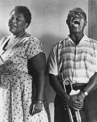 ella fitzgerald smiles and looks at the camera while wearing a short sleeve patterned dress, louis armstrong stands to the right with a wide mouth smile as he holds his trumpet, he wears a short sleeve button up shirt with pants and thick framed glasses