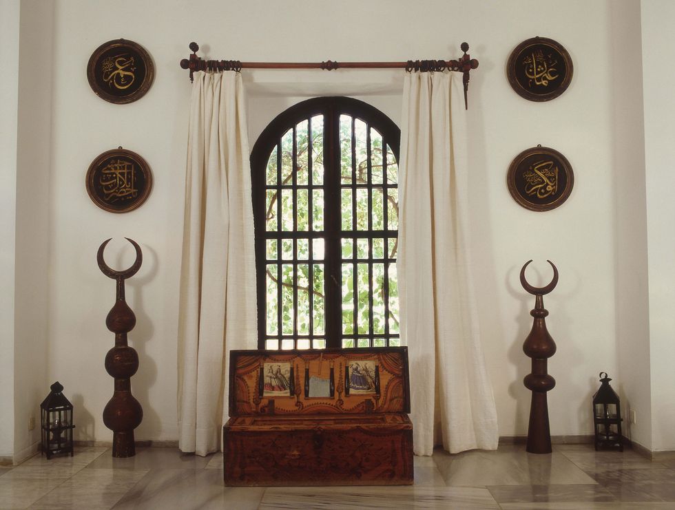 a white painted room with a tall arched window and a windowseat in front and shields on the wall flanking the window