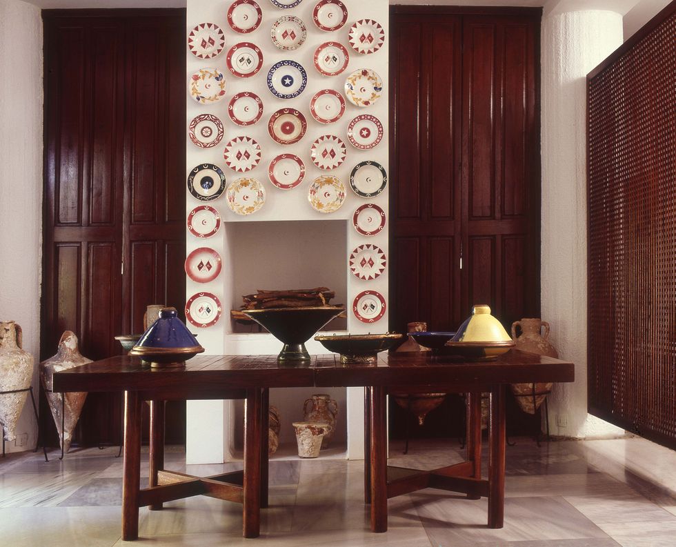 a white washed wall over a simple fireplace on which there are colored plates hung and in front a heavy wood table with objects and stone figures on the floor next to everything