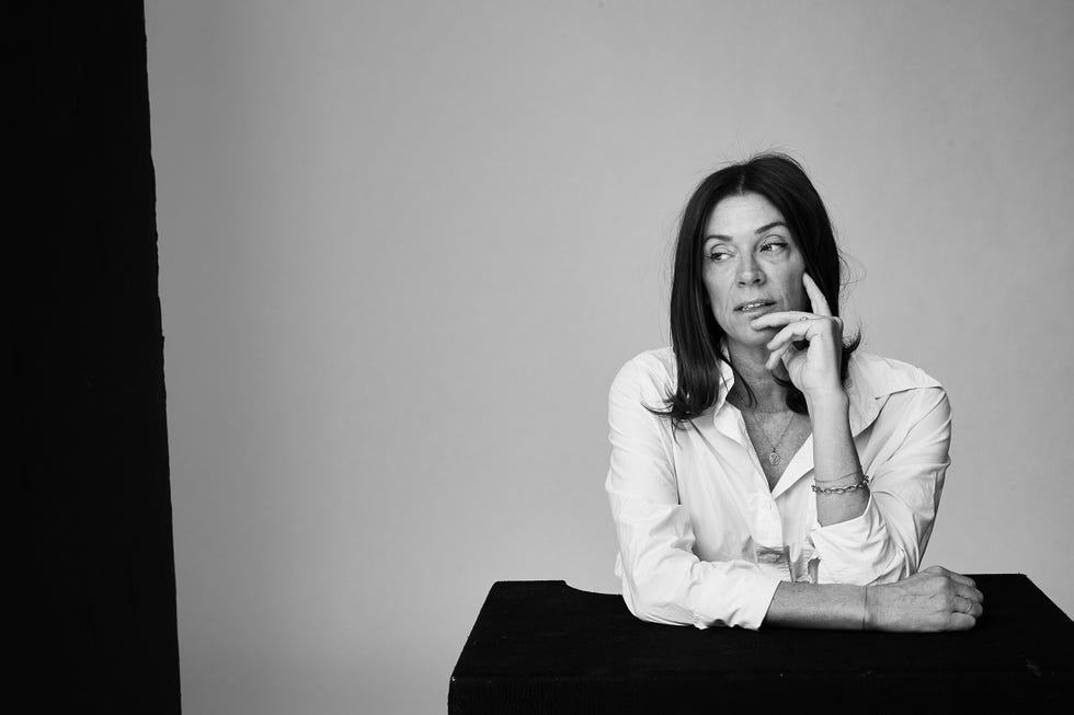 a person is seated at a black table leaning slightly forward with their chin resting on their hand they are wearing a white button up shirt which is slightly unbuttoned at the collar their hair is long and dark and the background features a gradient from light gray to a darker shade the table surface is matte contrasting with the smooth fabric of the shirt