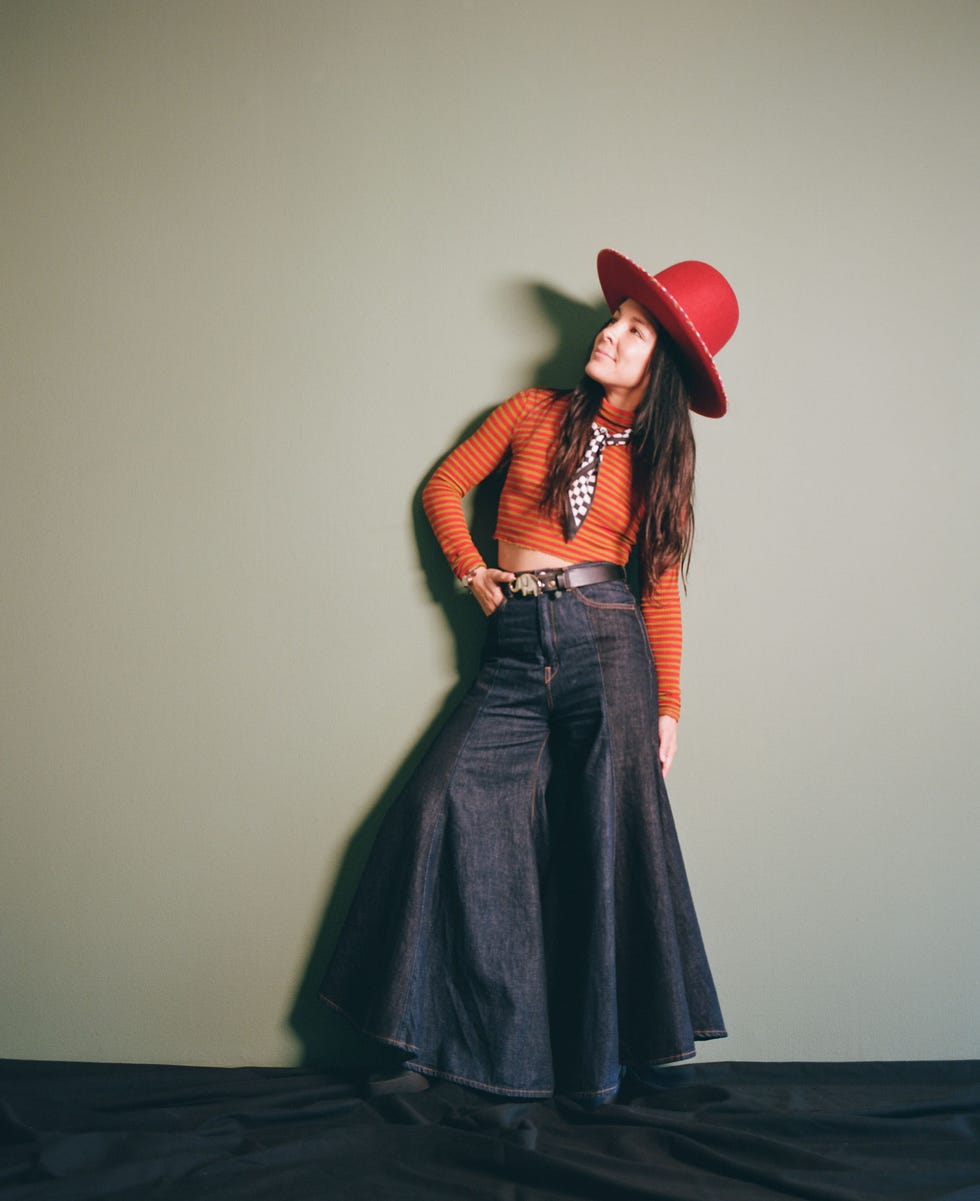 a person styled in a fashionable outfit with a large red hat against a plain green background