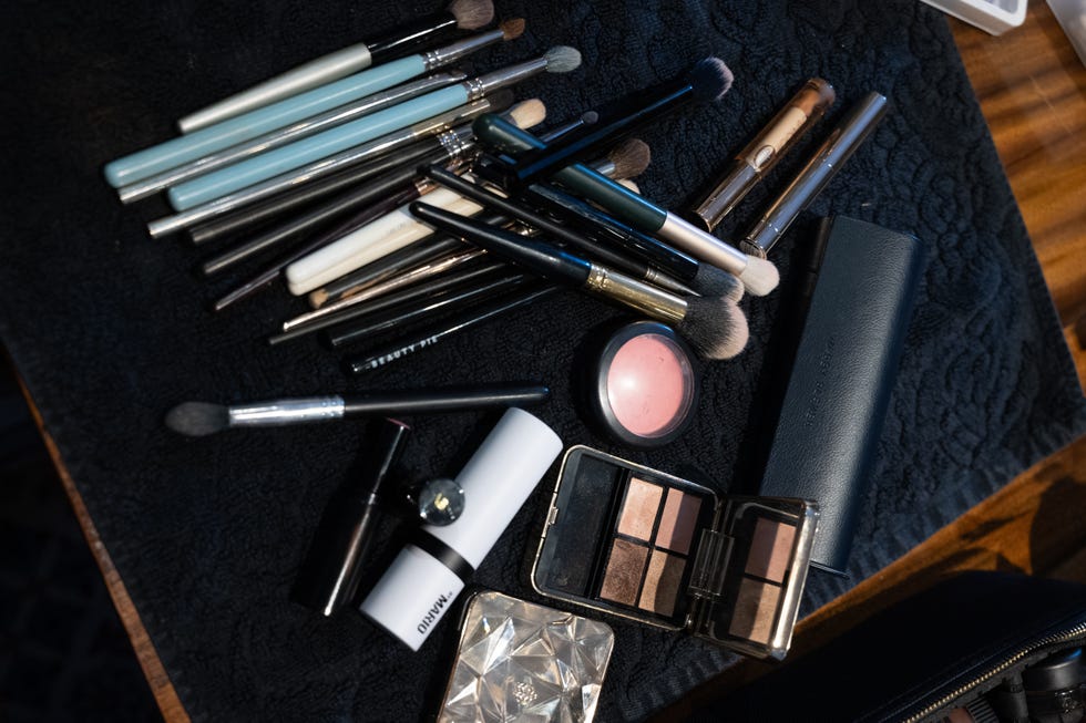 the scene features an assortment of makeup brushes, some with colorful handles and others in black, scattered across a dark textured mat several cosmetic items are also present, including a round blush compact, a small palette with multiple eyeshadow shades, and various containers for lip products the arrangement suggests an active makeup application or organization process