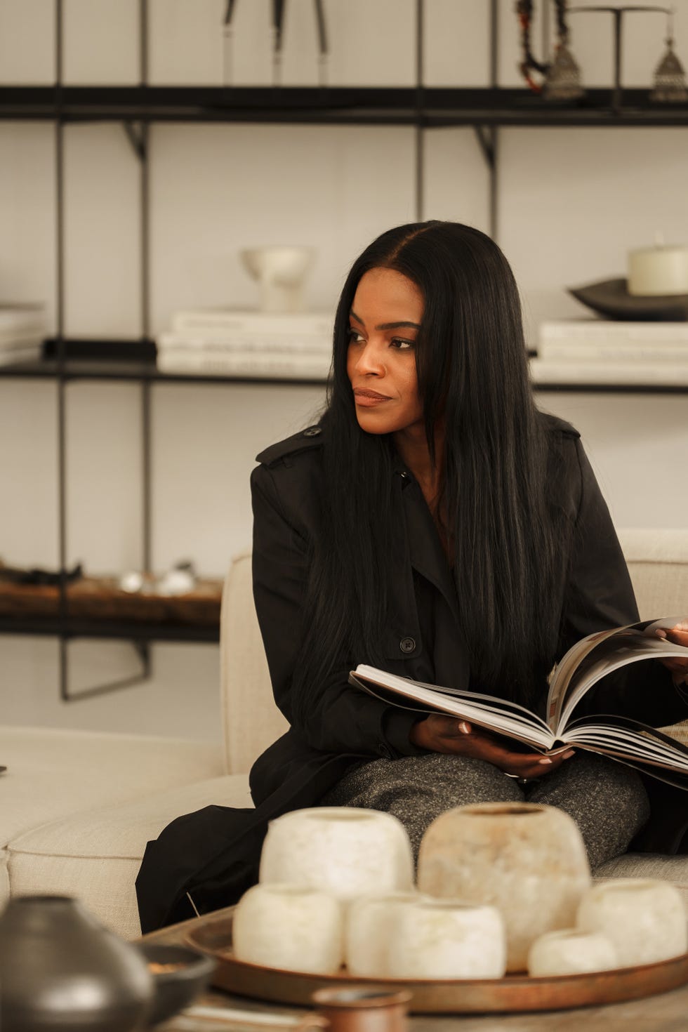miminat leafing through a book sat on a white sofa with black steel shelves behind her