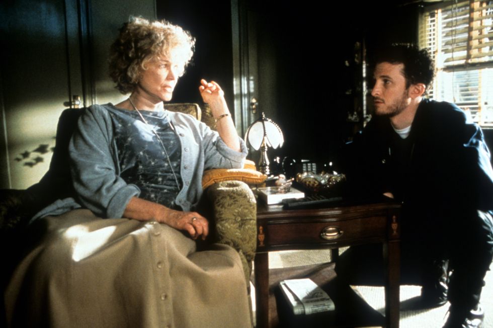 ellen burstyn in character for requiem for a dream, she sits in an arm chair and looks to the right at a man who is crouching nearby and looking at her, burstyn has a blue shirt and cardigan with a long camel skirt