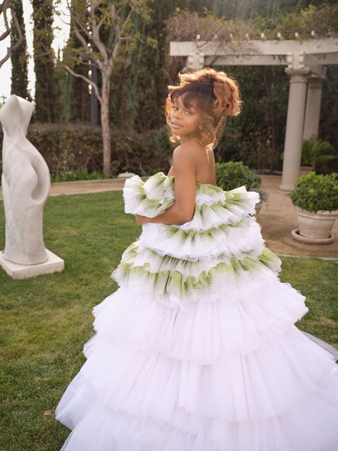 marsai martin stands in a garden near a marble figurative sculpture wearing a tiered white and green dress