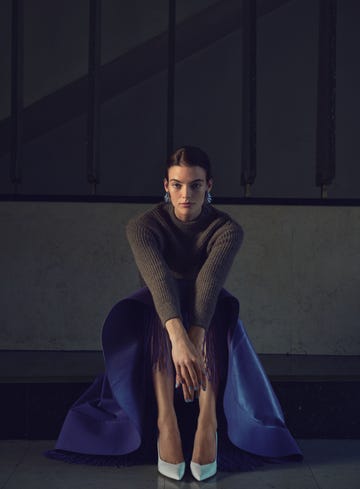 A seated figure wearing a gray sweater and a purple fringed skirt with white heels