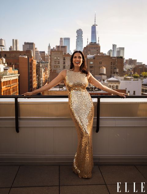 padma lakshmi stands on a balcony overlooking nyc, wearing a gold sequined gown