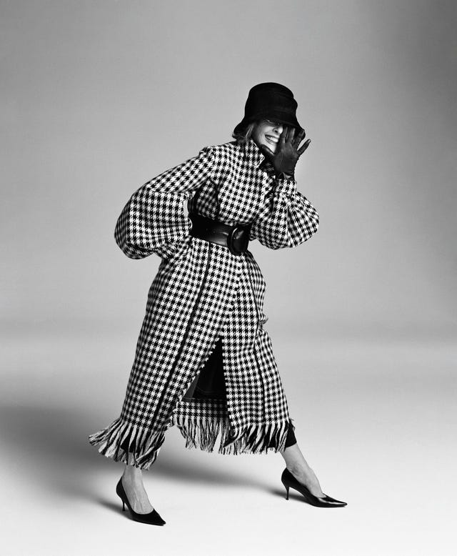 a woman in a houndstooth coat and black hat, raising her hand to her face