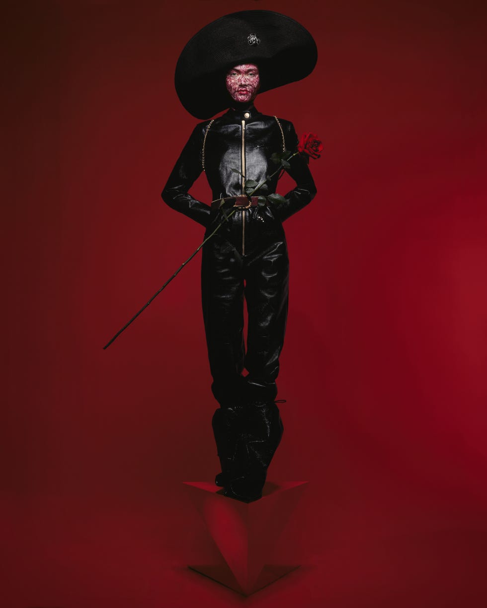 a woman in a black jumpsuit and hat standing against a red background