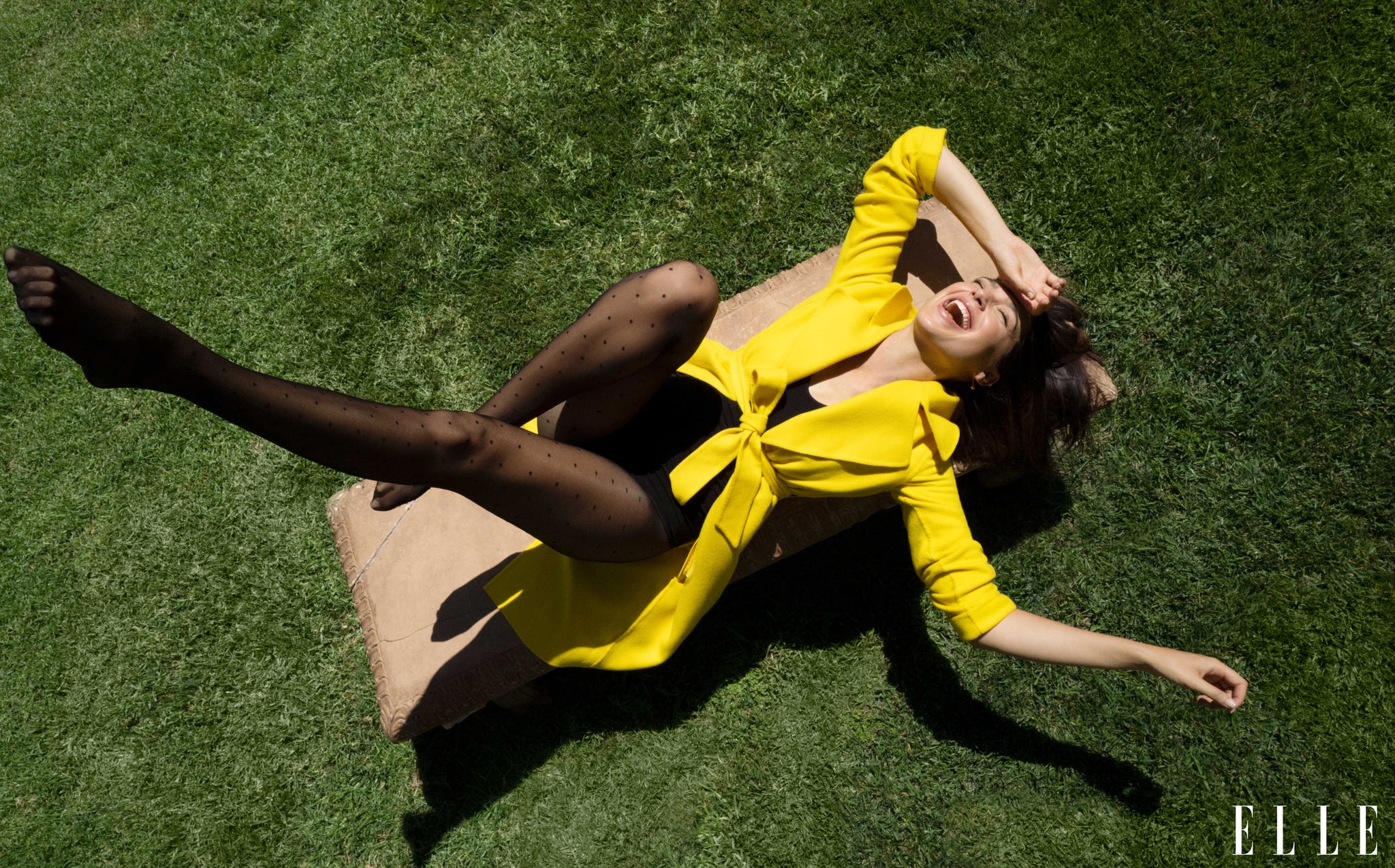 gal gadot lies on a bench on the grass, laughing, in a yellow trench coat and tights