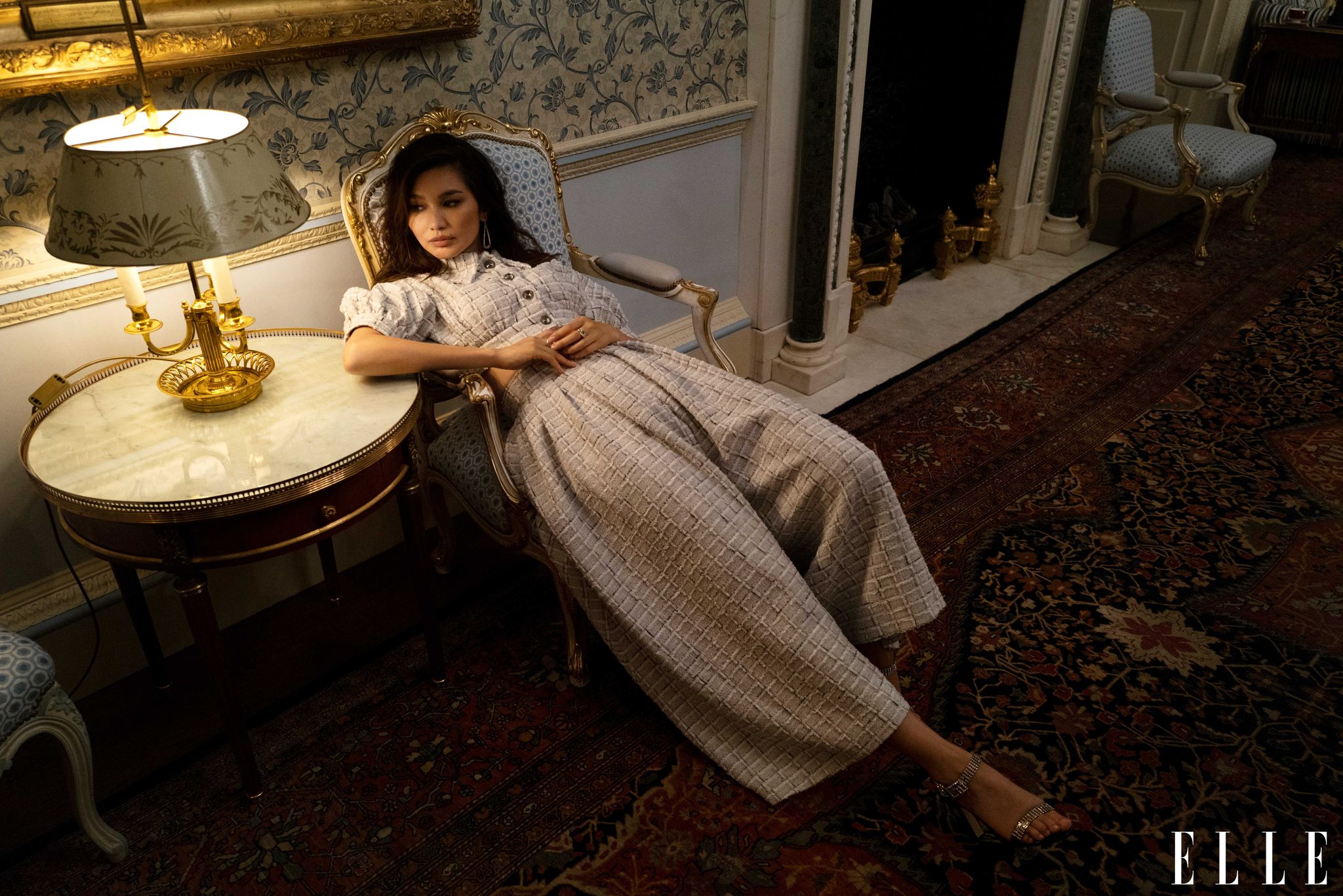 gemma chan slouches in an armchair in an ornate hallway