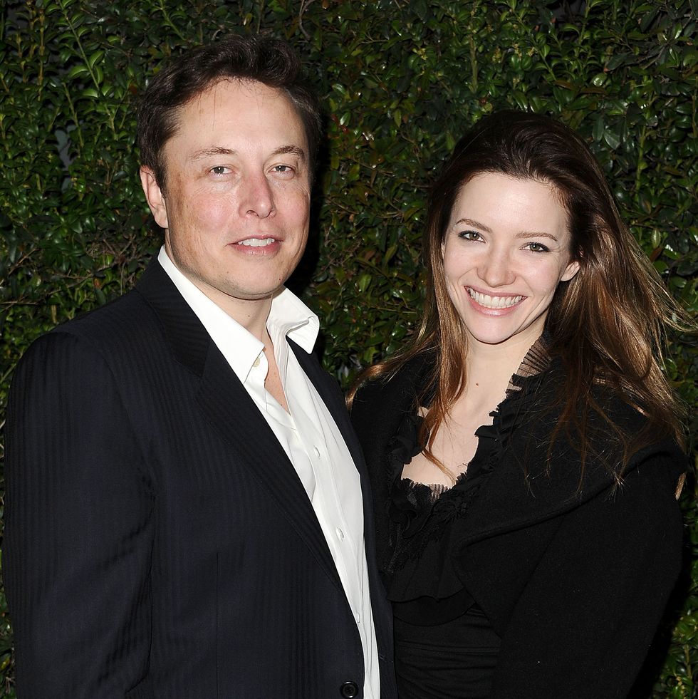 elon musk and talulah riley stand together and smile for photos, he wears a suit jacket and white collared shirt, she wears a black coat and outfit