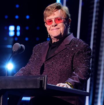 elton john smiles at the camera from behind a podium with a microphone, he wears a purple bejeweled suit jacket, black collared shirt, and pink tinted glasses