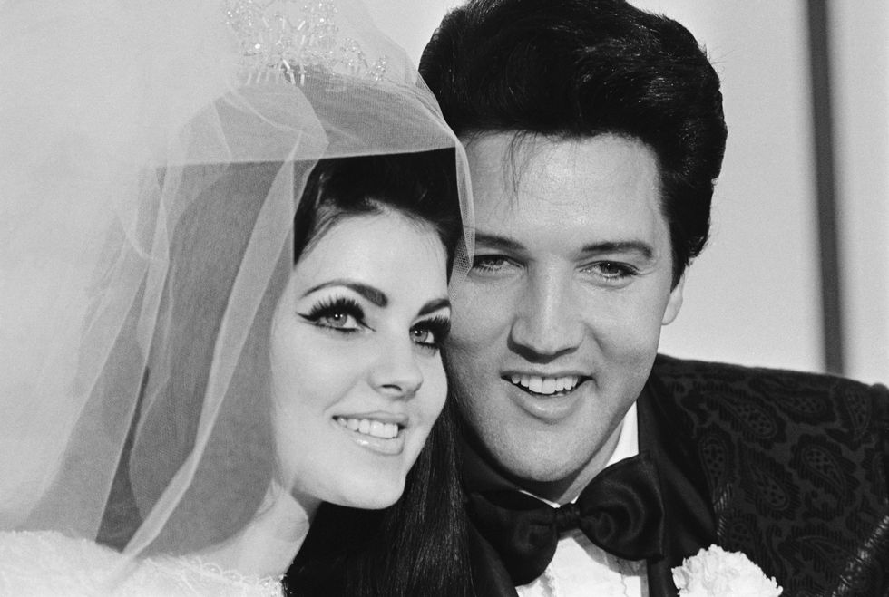 priscilla presley wearing her wedding dress and veil and smiling for a photo with elvis, also smiling and wearing a black tuxedo
