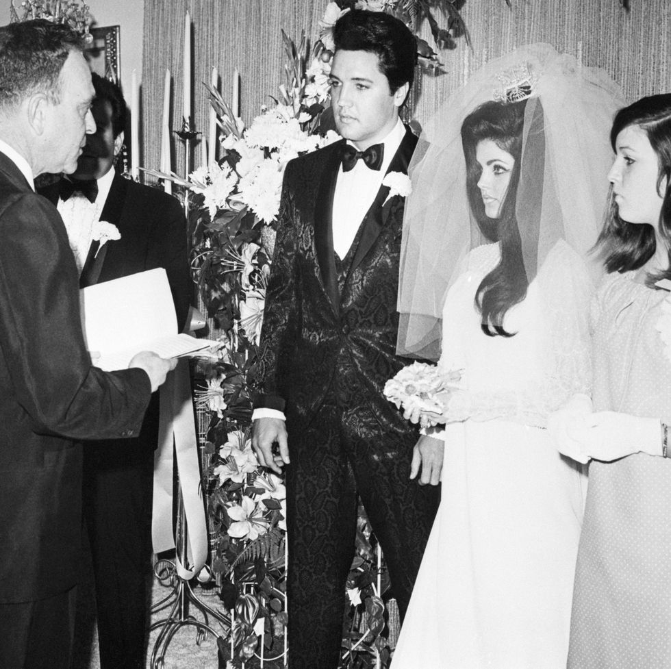 elvis and priscilla presley stand together and face a man who reads from a book he holds, elvis wears a black patterned tuxedo with a white carnation on his lapel, priscilla wears a white wedding dress and veil and holds a small flower bouquet, another woman in a dress stands on the right, a large flower arrangement is in the background