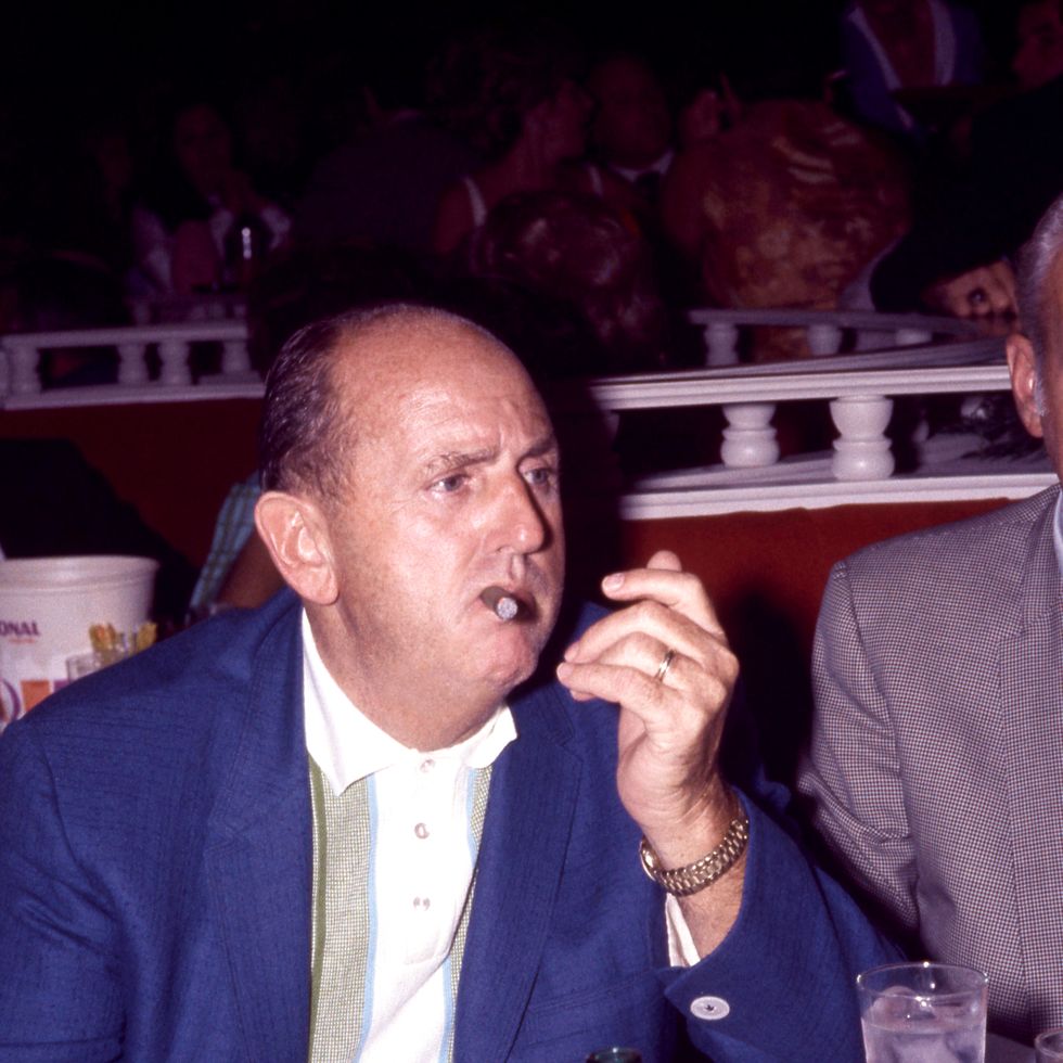 a man in a blue suit jacket and polo shirt smokes a cigar while sitting at a dining table in a dark room, he looks to the right and leans against the table on one elbow