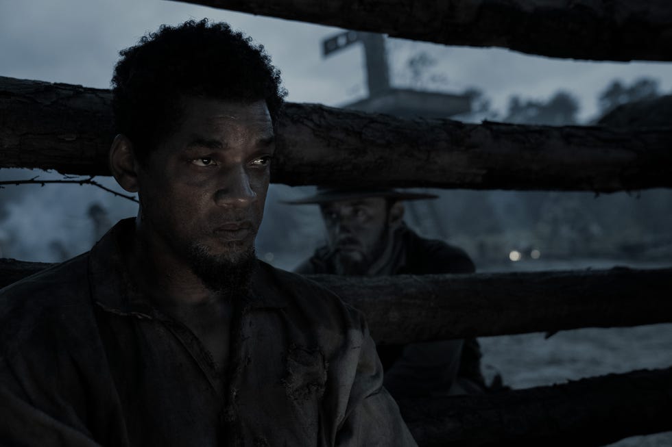 The scene shows two people, one prominently positioned in the foreground and another partially visible in the background. They are surrounded by large, rough-hewn tree trunks that form a barrier. The background is dark and moody, suggesting a cloudy or dim environment