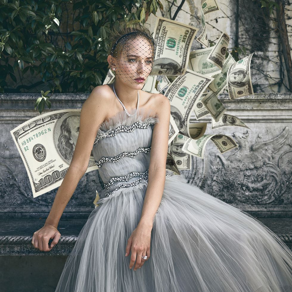 bride sitting on a stone bench in a garden in a dove grey dress and accessorized with diamond jewelry, designed with dollar bills scattered around her