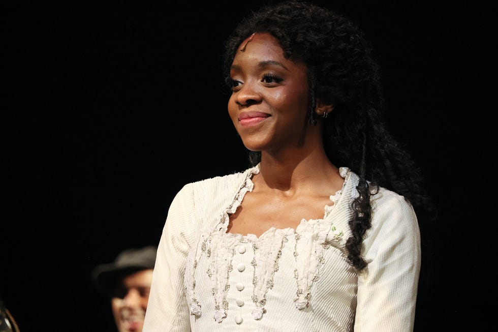 Emilie Kouatchou in a 2023 “Phantom of the Opera” performance emilie kouatchou wearing a white dress and smiling on a darkened stage