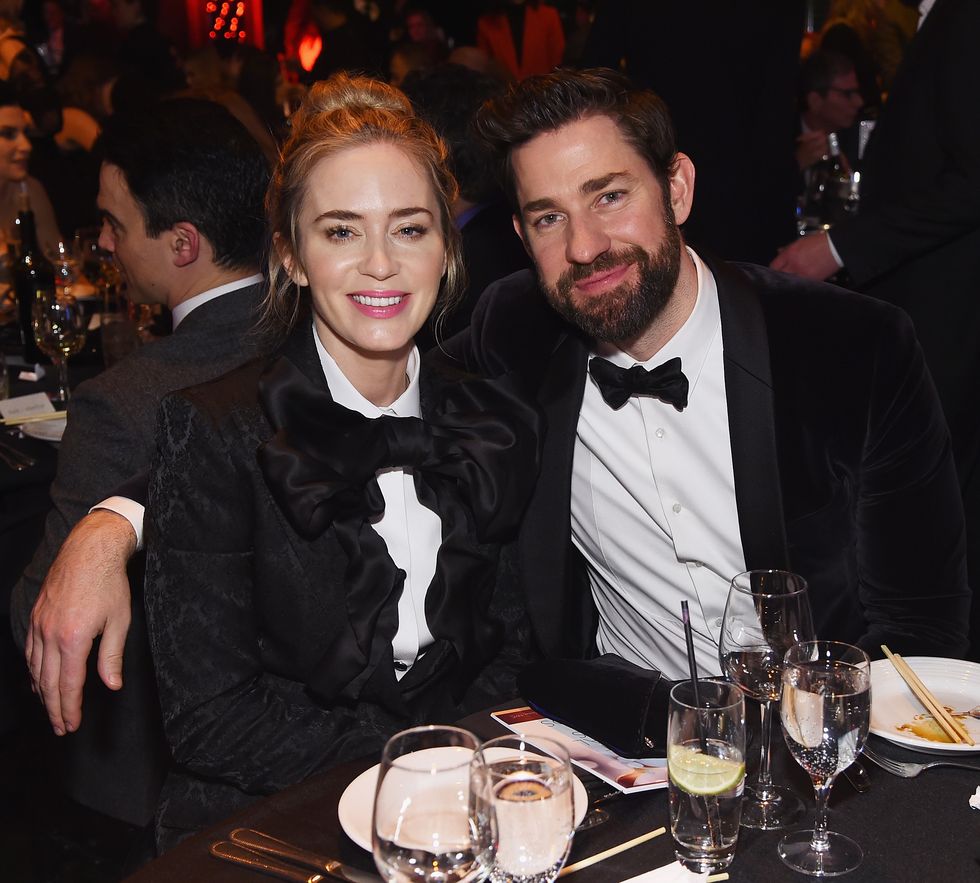 emily blunt and john krasinski sit at a table and smile for a photo, both wear black bow ties with white collared shirts, she also wears a black jacket, he has his arm around the back of her chair